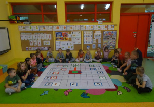 Grupa dzieci siedzi wokół maty ułożonej na dywanie. Na macie ułożony jest zegar z tabliczek z cyframi oraz wskazówek papierowych. Obok każdej godziny leży obrazek informujący co dzieci robią co dzień o danej godzinie.